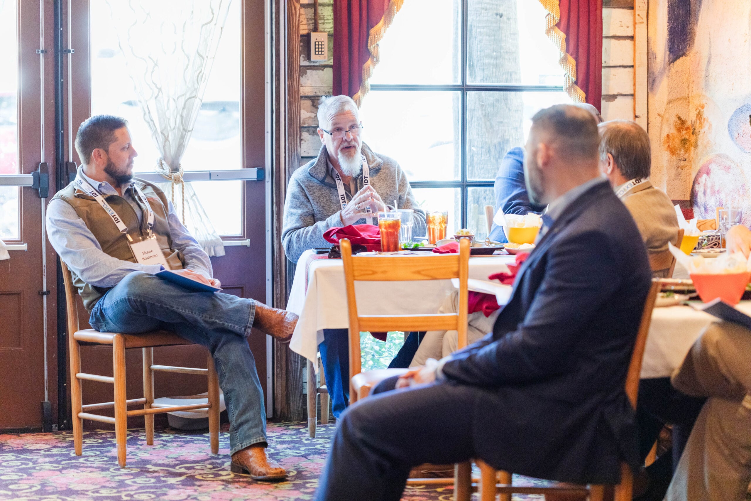 A group of men sit around a dining table in a banquet room. They are in discussion and daylight comes in through a curtained window.
